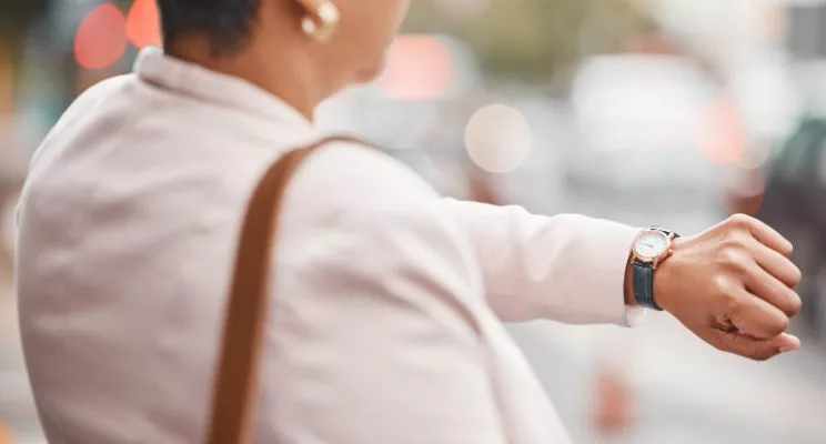 A woman wearing a business blazer stands on a blurred busy street, looking at her watch, conveying a sense of time management and the hustle and bustle of the urban environment.